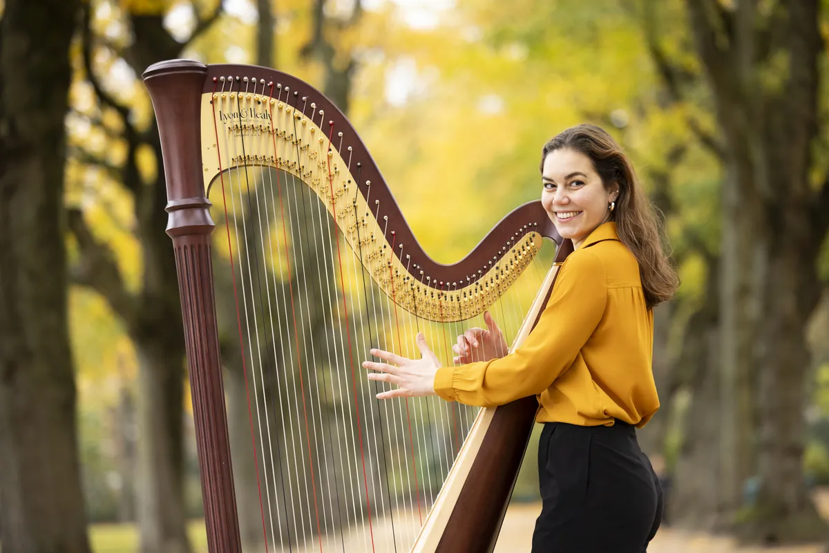 Eleonora Congiu with both hands on the strings of her pedal harp outdoors among autumn trees