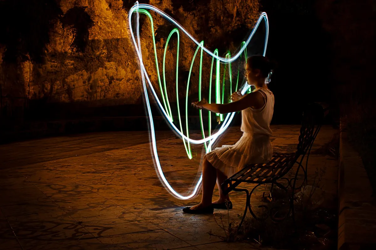 A harp drawn in light against a floodlit wall at night, a player seated beside it