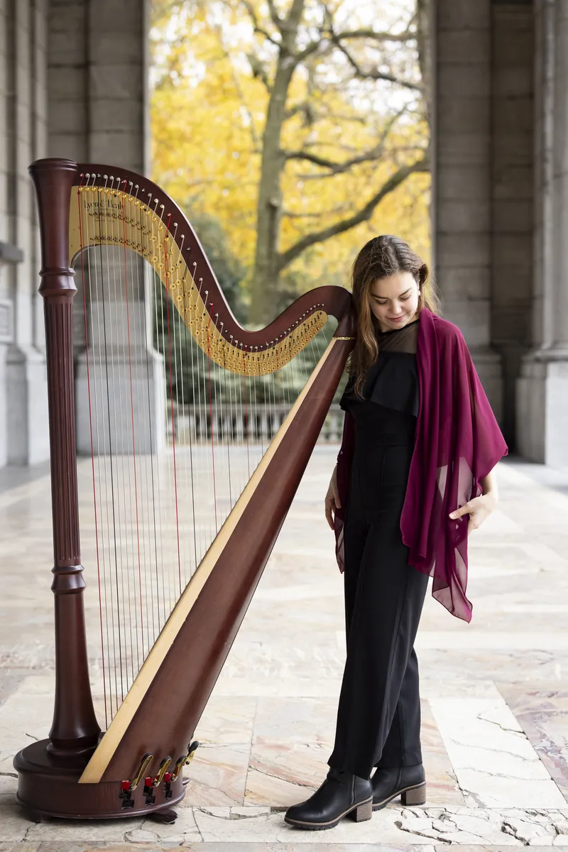 Eleonora Congiu in black with a burgundy shawl, leaning against the neck of her pedal harp in a colonnade