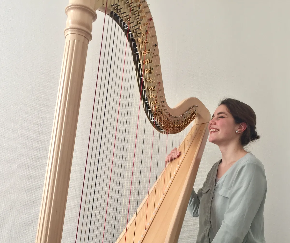 Eleonora Congiu laughing beside her pedal harp in daylight against a plain wall
