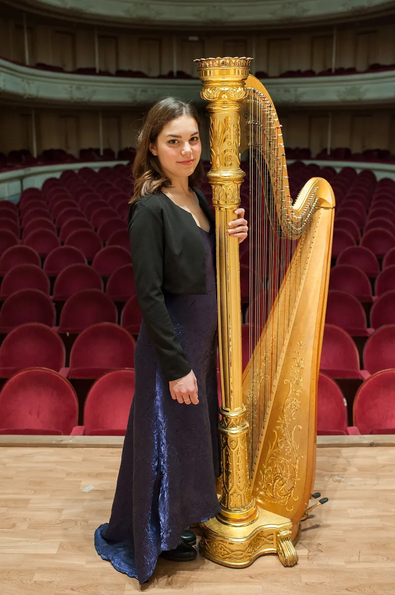 Eleonora Congiu standing beside a gilded pedal harp in an empty theatre, red seats behind her
