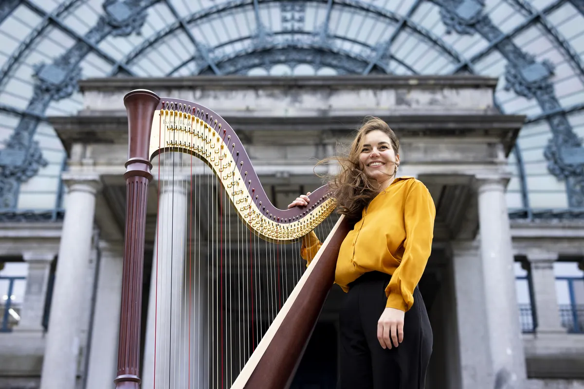 Eleonora Congiu standing with her pedal harp under a glazed iron and stone portico, looking up into the light