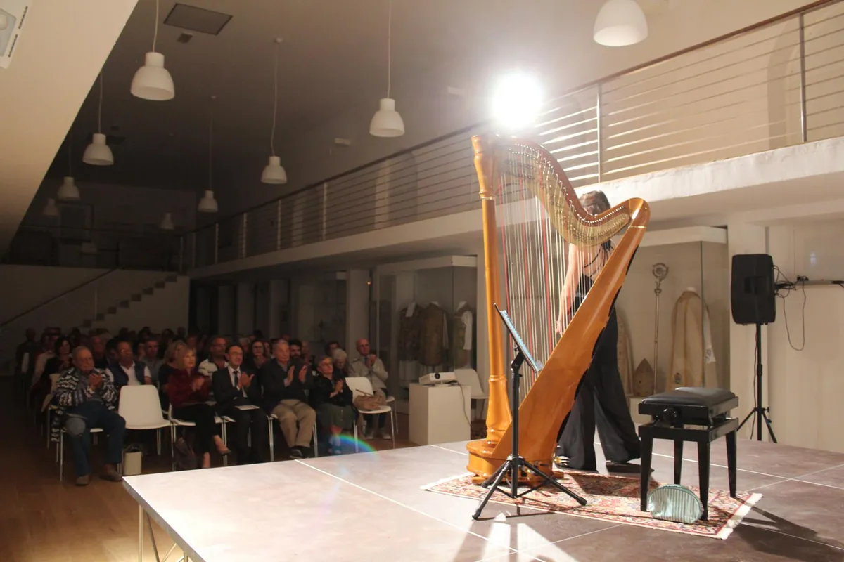 A lit pedal harp on a museum stage with the audience seated in rows beyond it