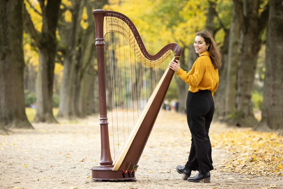 Eleonora Congiu standing at her pedal harp on a leaf-covered path, looking back over her shoulder
